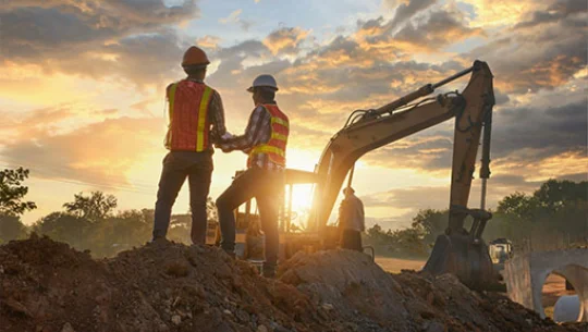 A construction crew stands on a mound of earth at a job site, with heavy machinery in the background.